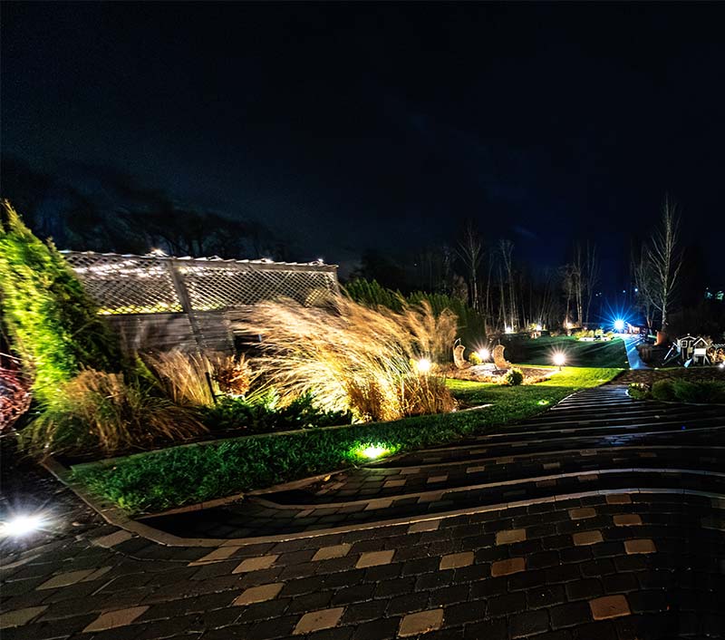 A curved paver path descends past low lights, grasses, shrubs, and a lit lattice fence at a private residence at night