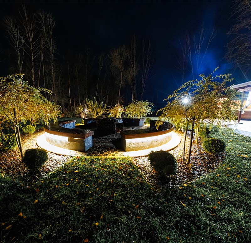 A ring seating area glows at night with underlighting around a brick firepit. Small trees and shrubs frame wet grass and leaves.